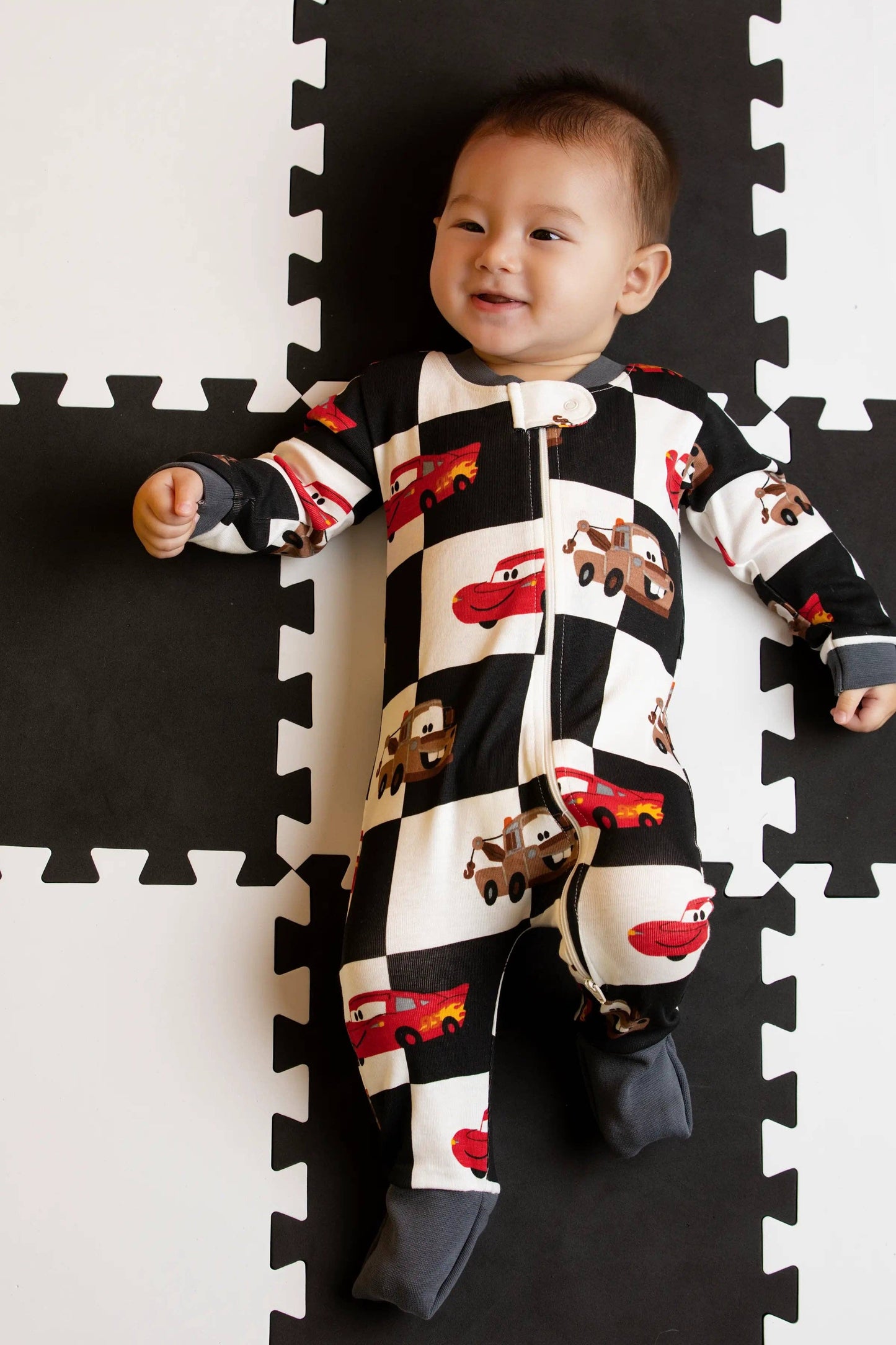 Baby wearing a black and white checkered onesie with vehicle patterns on a foam puzzle mat.