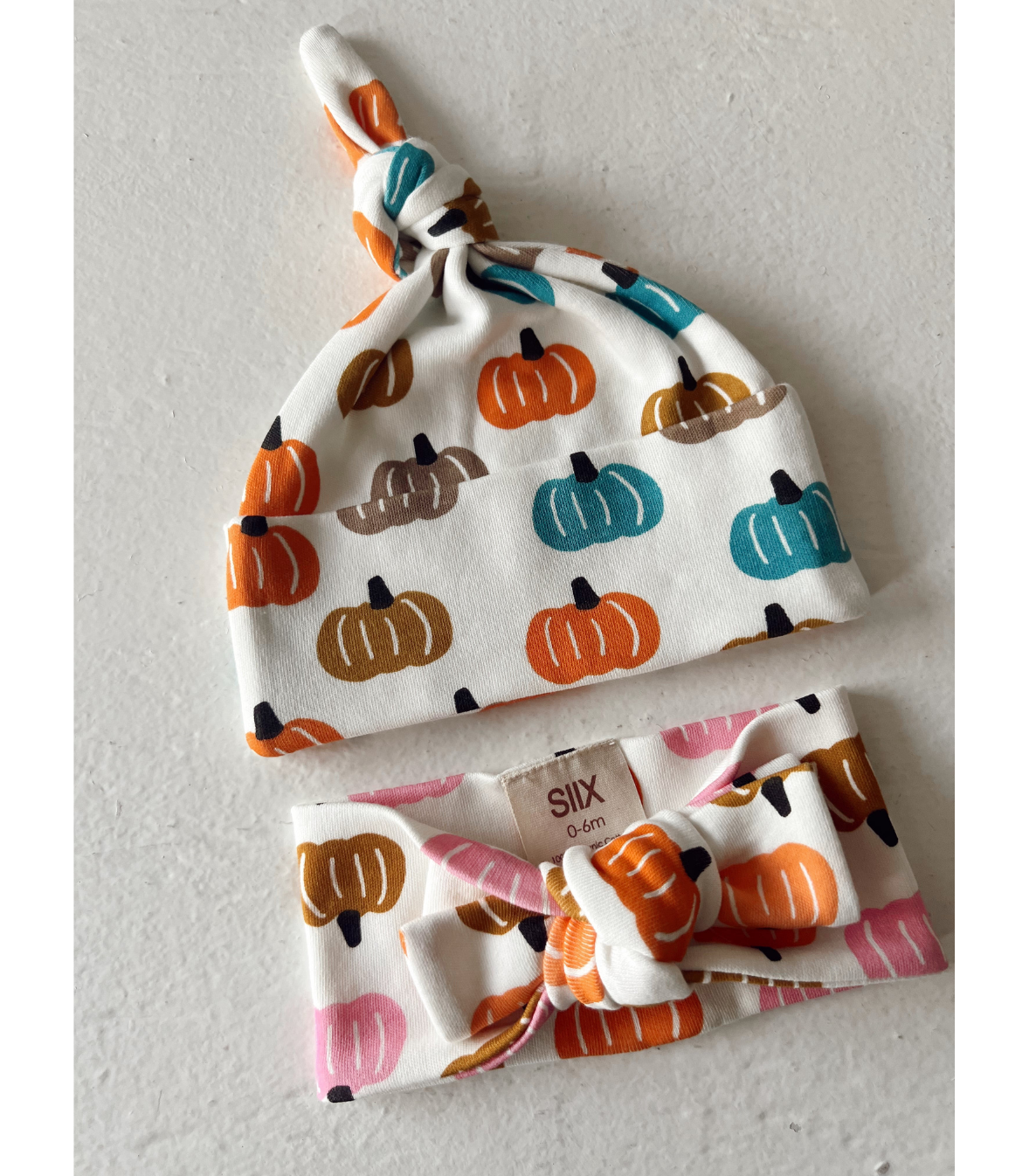 A baby hat and matching headband set by Slix Baby, featuring a white fabric print of orange, brown, and teal pumpkins. The hat has a knotted top, and the headband has a large bow.