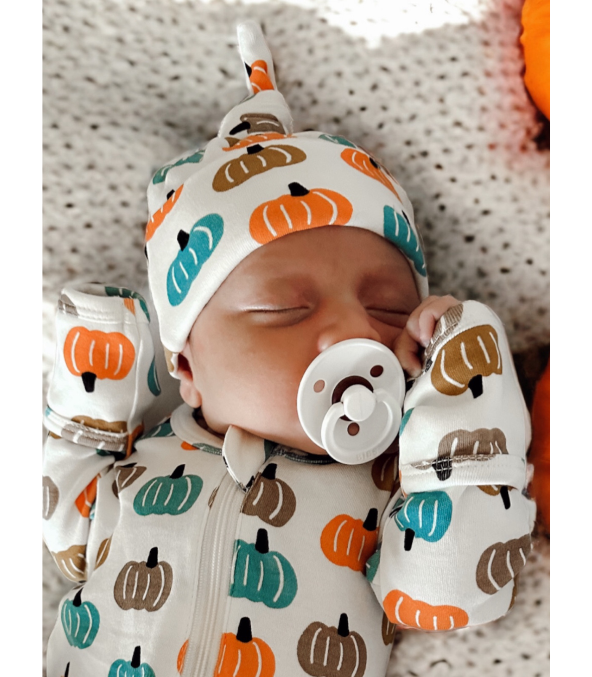 A sleeping newborn baby wearing a white zip-up onesie and matching beanie hat with an orange, teal, and brown pumpkin print, holding a pacifier.