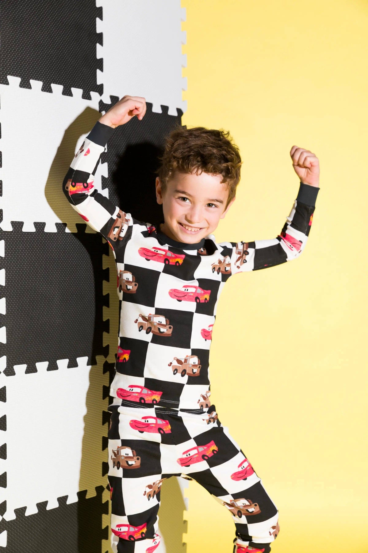 Child wearing a checkered pajama set with car design on a yellow and black puzzle mat.