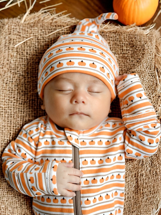 A sleeping newborn baby wearing an orange and white striped knit one-piece outfit with a pumpkin print, lying on a burlap blanket with a small orange pumpkin in the background.