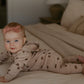 A baby wearing a beige hooded one-piece outfit with brown leaf prints and a matching pink bow headband, lying on a bed in a softly lit bedroom.