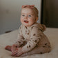 A smiling baby sitting on a bed wearing a beige hooded one-piece outfit with brown acorn prints and a matching pink bow headband.