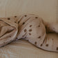 A close-up of a baby's legs and torso wearing beige pajamas with a brown pinecone pattern, lying on a textured beige blanket.