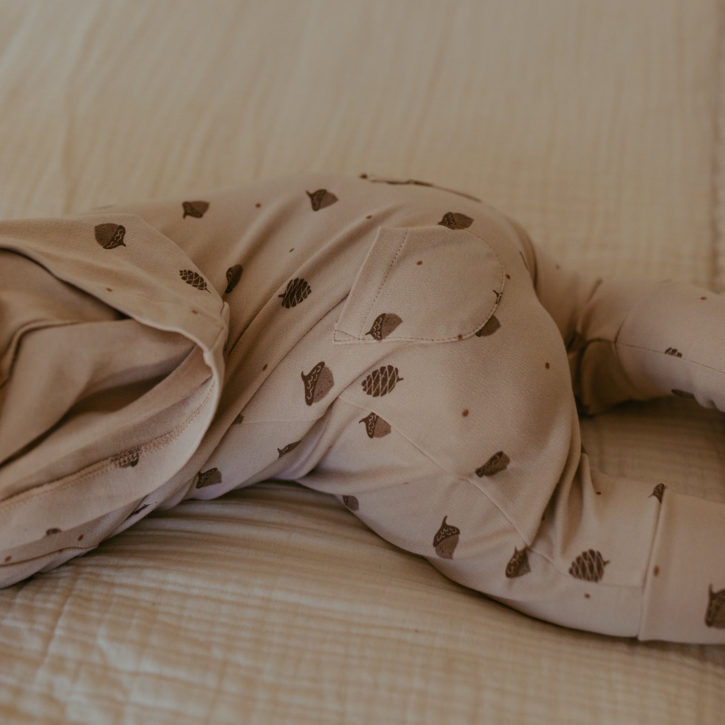 A close-up of a baby's legs and torso wearing beige pajamas with a brown pinecone pattern, lying on a textured beige blanket.