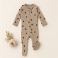 A baby footed sleeper in a warm beige color, decorated with a pattern of brown acorns and pinecones, laid flat on a light cream background next to a small decorative pumpkin.