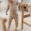 A baby wearing a beige footed zip-up sleeper with dark brown pinecone prints, standing on a wooden floor and holding onto a wooden chair.