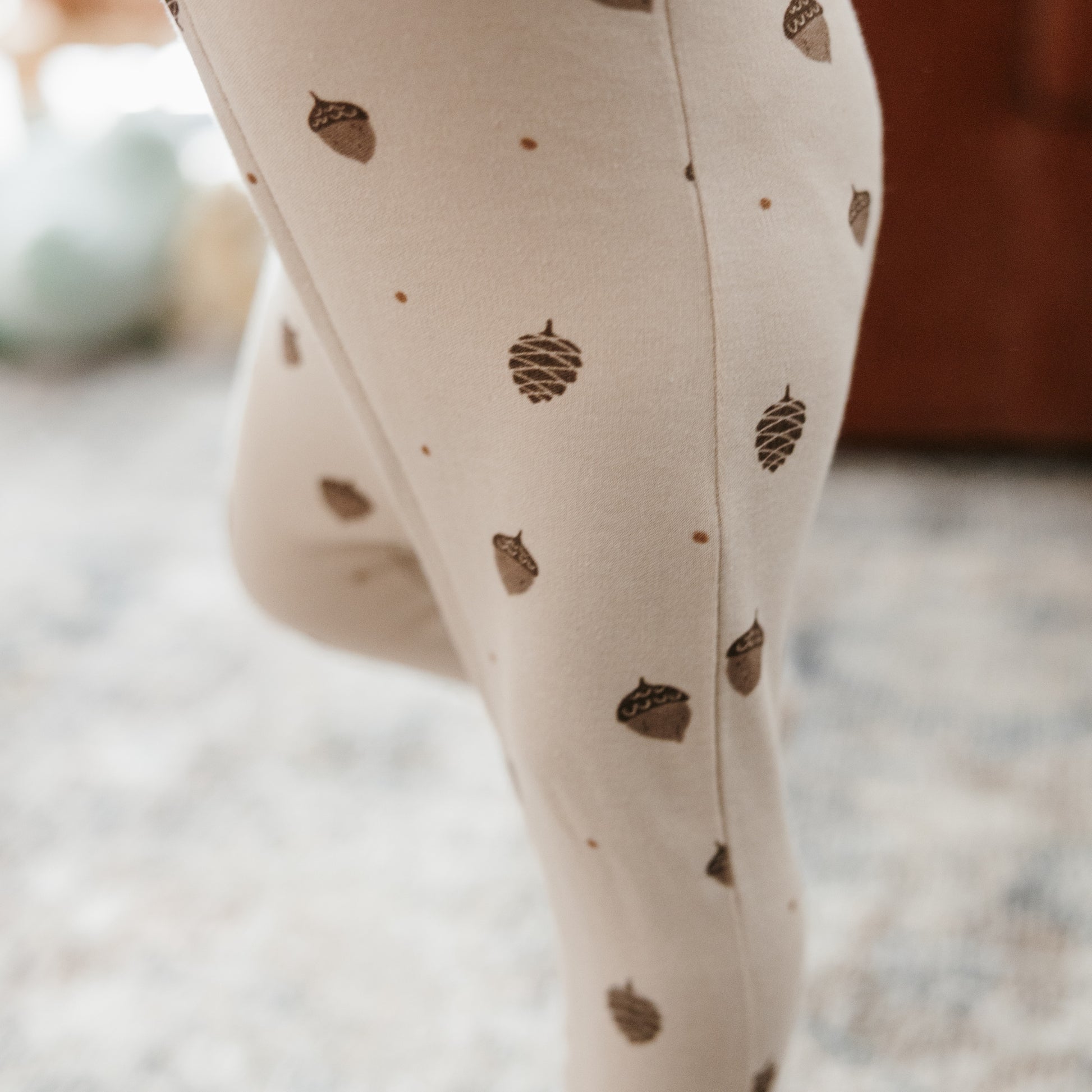 Close-up of a person wearing cream leggings with a brown acorn and pinecone pattern, standing on a blurred indoor floor.