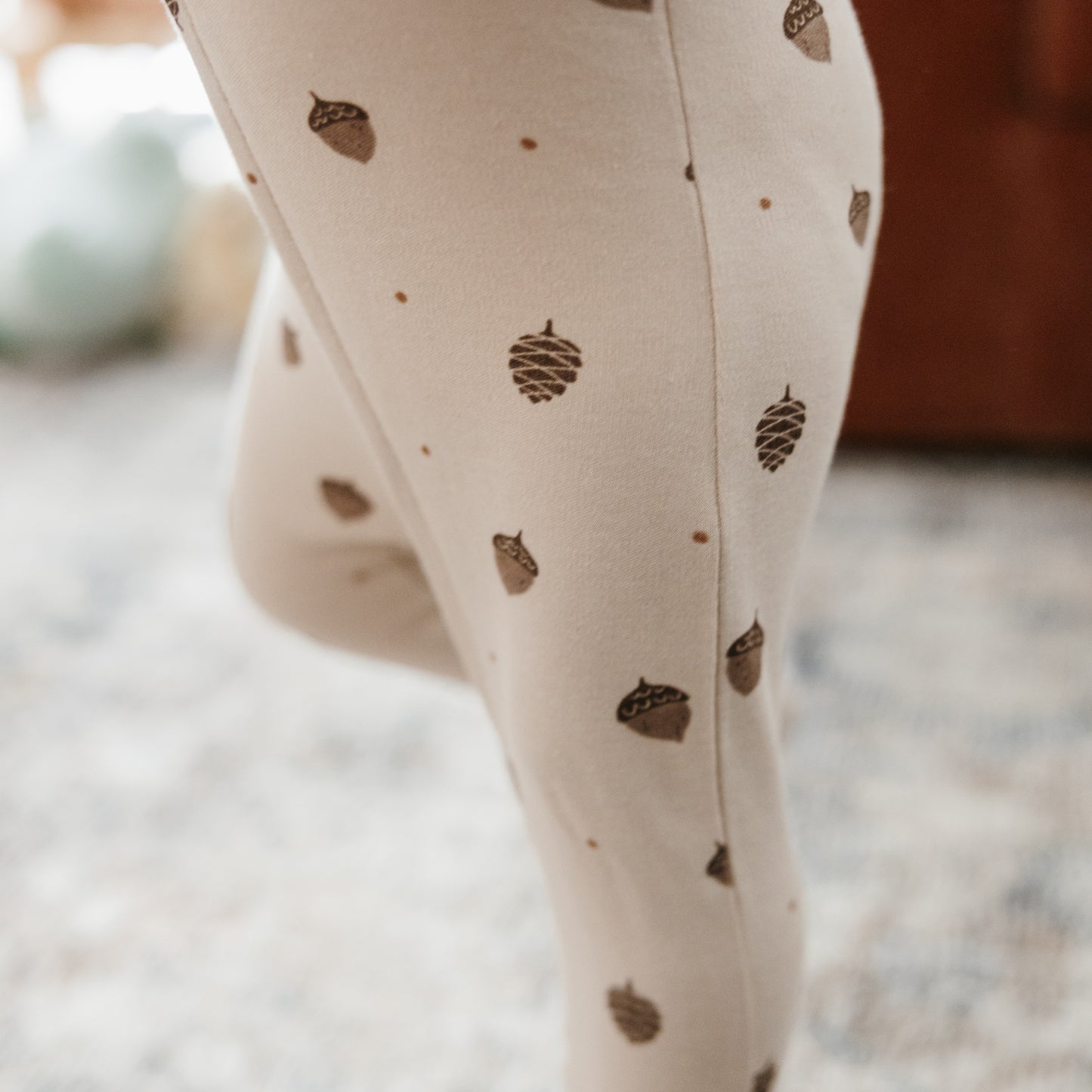 Close-up of a person wearing cream leggings with a brown acorn and pinecone pattern, standing on a blurred indoor floor.