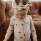 A toddler wearing a cream-colored long-sleeve footless pajama top with brown polka dots and a matching beige fabric bow headband, standing in front of a brown leather couch.