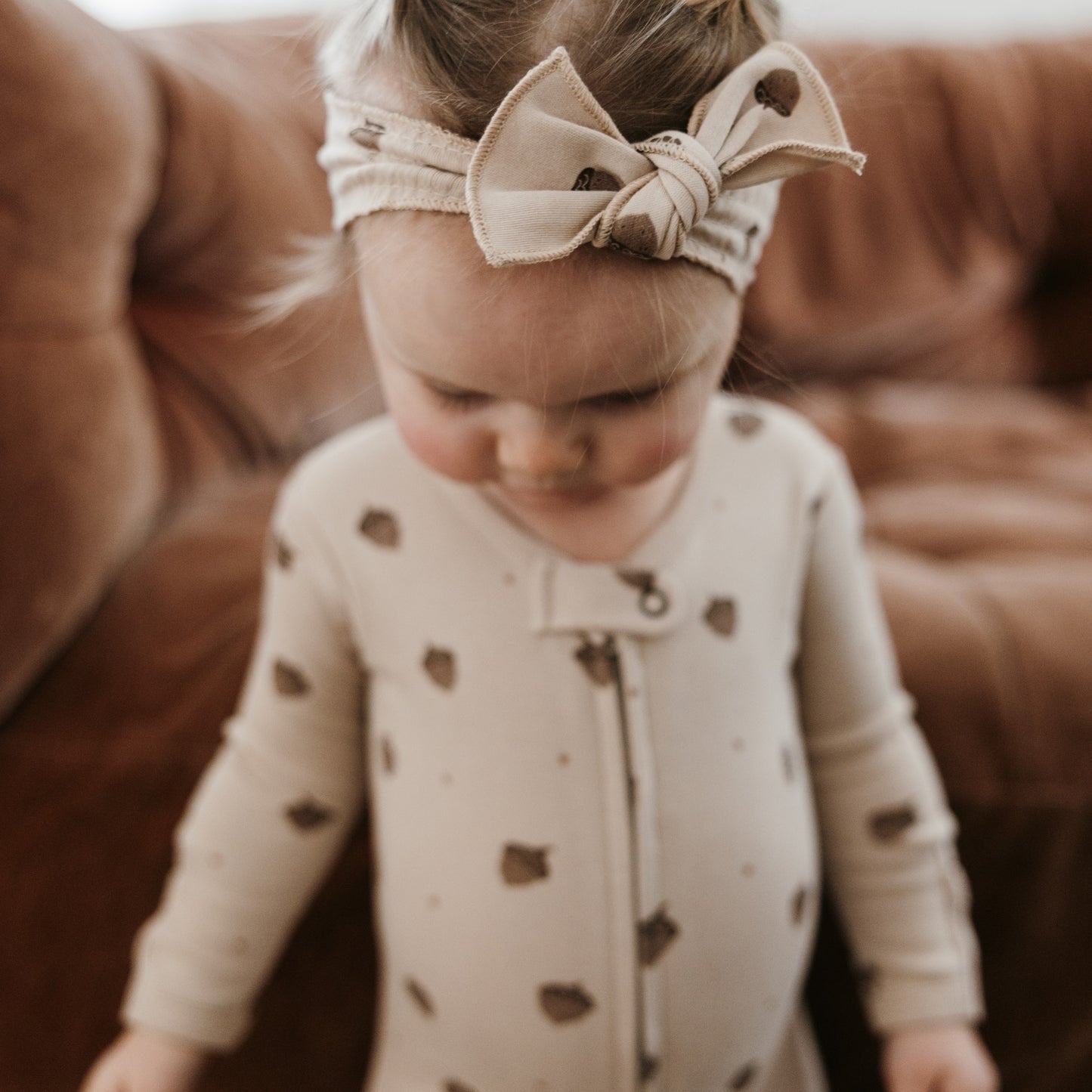 A toddler wearing a cream-colored long-sleeve footless pajama top with brown polka dots and a matching beige fabric bow headband, standing in front of a brown leather couch.