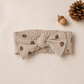 A baby headband made of soft, textured beige fabric with a printed pattern of brown acorns and pinecones, featuring a large bow on the front. The headband is displayed flat on a neutral beige background with decorative props, including a pinecone and acorn.