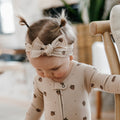 A baby wearing a cream-colored long-sleeve zip-up onesie with brown acorn and pinecone prints, paired with a matching bow headband, standing next to a wooden chair.