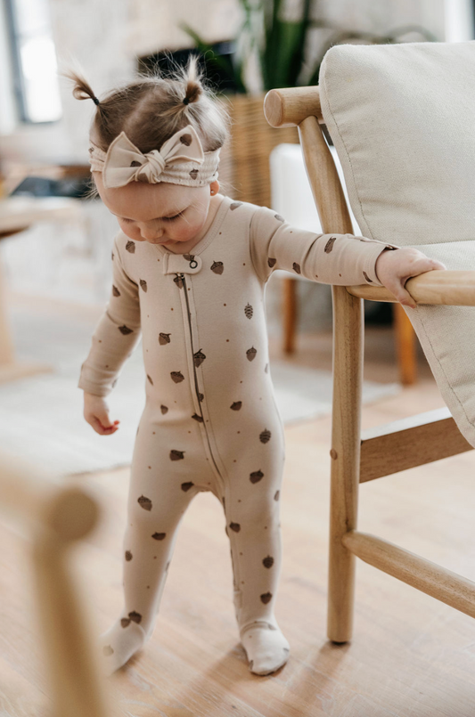 A toddler girl wearing a beige footed one-piece sleeper with brown heart prints and a matching bow headband, standing indoors next to a wooden chair.