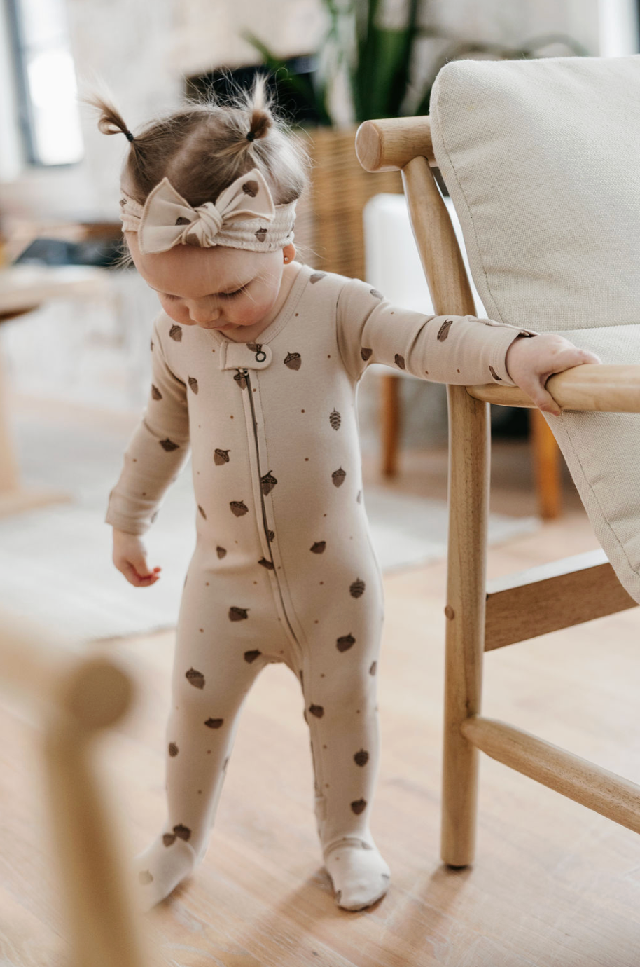 A toddler girl wearing a beige footed one-piece sleeper with brown heart prints and a matching bow headband, standing indoors next to a wooden chair.