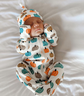 A sleeping newborn baby wearing a white footed onesie and matching hat with an orange, teal, and brown pumpkin print, lying on white bedding.