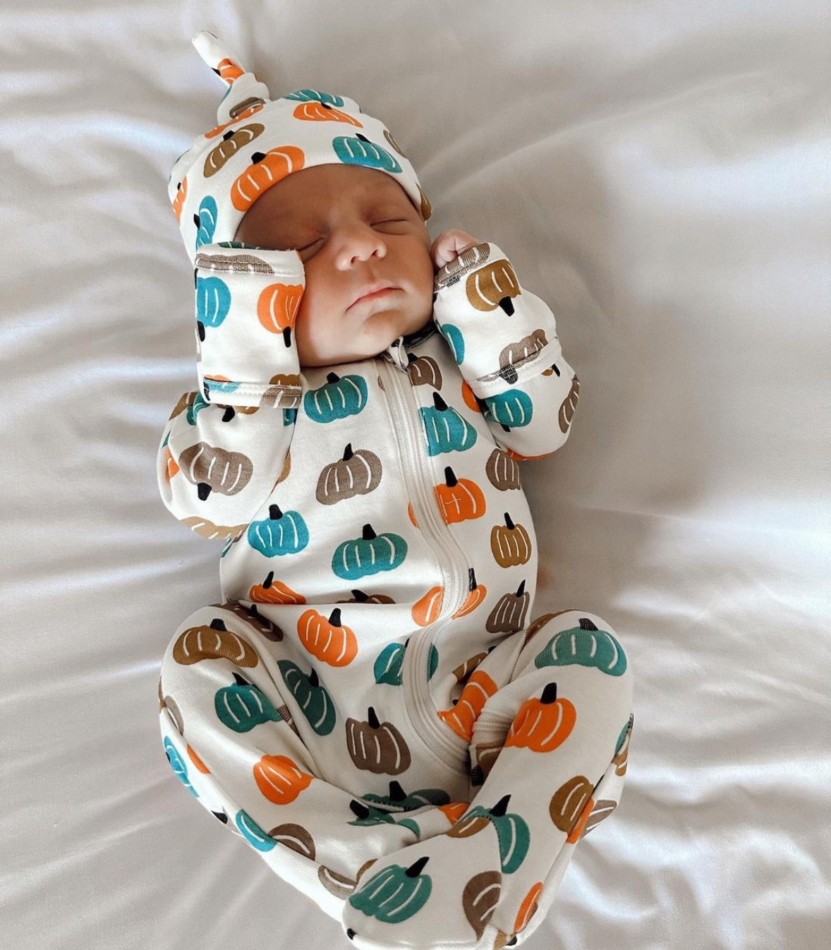 A sleeping newborn baby wearing a white footed onesie and matching hat with an orange, teal, and brown pumpkin print, lying on white bedding.